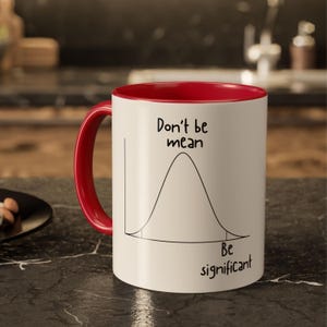 Puede incluir: Una taza de cerámica blanca con interior y asa de color rojo brillante. El exterior presenta un dibujo lineal negro de una curva de campana con el texto "Don't be mean" sobre el pico y "Be significant" escrito bajo la curva a la derecha.