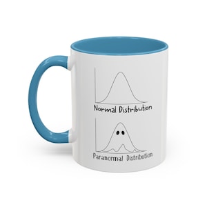 May include: White ceramic mug with a blue rim and handle. The mug features a graphic of two bell curves, one labeled "Normal Distribution" and the other labeled "Paranormal Distribution". The "Paranormal Distribution" curve is shaped like a ghost.