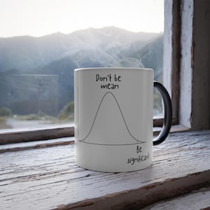 May include: White ceramic mug with a black handle, featuring a black bell curve graphic and the phrases "Don't be mean" and "Be significant" in black text. Steam rises from the mug, suggesting a hot beverage.
