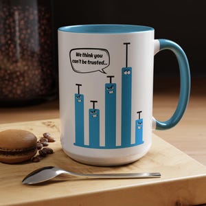 May include: White ceramic mug with a blue interior and handle. The mug features a graphic of blue bar graphs with angry faces and the text "We think you can't be trusted..." in a speech bubble. A spoon and a macaron are on the wooden surface.