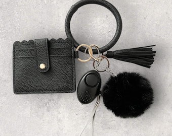 May include: A black leather card holder with a scalloped edge, a black faux fur keychain, a black silicone ring keychain with a black tassel, and a black personal alarm keychain.