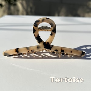May include: Tortoise hair claw clip. This claw clip features a tortoiseshell pattern in shades of beige and dark brown. The clip has a gold-colored spring mechanism and is designed to hold hair securely. The word "Tortoise" is printed at the bottom.