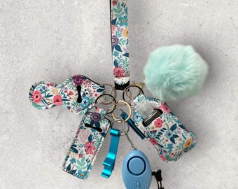 May include: A floral patterned keychain with a variety of accessories, including a hand sanitizer holder, a bottle opener, a personal alarm, and a fluffy pom-pom.