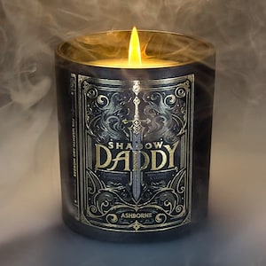 May include: A lit candle in a dark glass jar with a gold label. The label reads "SHADOW DADDY" with a sword graphic. The candle is surrounded by smoke, and the text "FOR WARMTH AND WONDER" is visible.