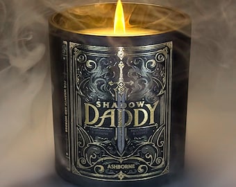 Puede incluir: Una vela encendida en un frasco de vidrio oscuro con una etiqueta dorada. La etiqueta dice "SHADOW DADDY" con un diseño de espada. La vela está rodeada de humo, y el texto "FOR WARMTH AND WONDER" es visible.