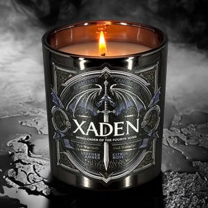 May include: Black and gold candle jar with a gothic design featuring a bat and sword. The label reads "Xaden, Wingleader of the Fourth Wing" and includes the scents "Leather, Amber, Citrus, Rose".