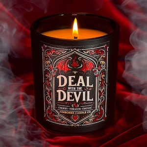 May include: Black candle with a red and gold label that says "Deal with the Devil" and "Cherry - Tobacco - Cognac" with a red dragon design. The candle is lit and there is smoke in the background.