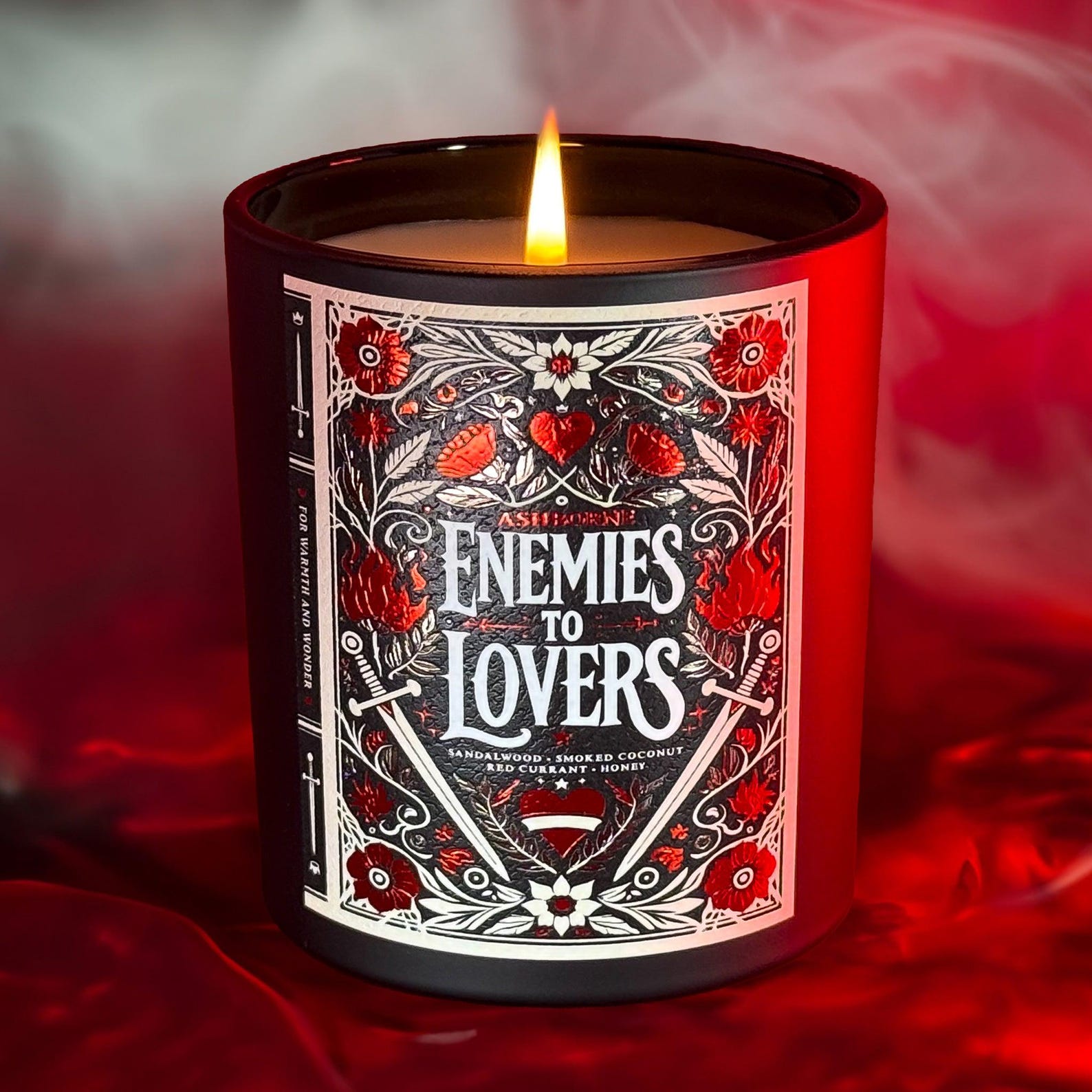 A lit candle with a decorative label that says "Enemies to Lovers" featuring floral and heart motifs, crossed swords, and a flame design.