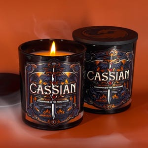 May include: Two black glass candles with the word "Cassian" in white lettering. One candle is lit, with a flame. The labels feature a sword, wings, and the words "Commander of the Night Court". The background is orange.