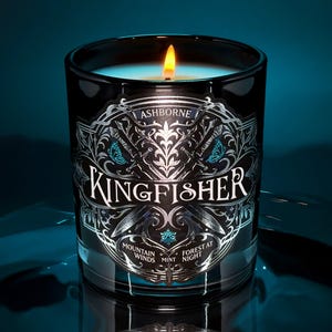May include: A lit black glass candle with a silver and teal label. The label reads "ASHBORNE KINGFISHER" with decorative elements and the phrases "Mountain Winds", "Mint", and "Forest at Night". The candle has a teal wax and a bright yellow flame.