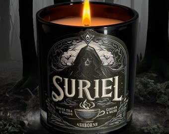 Puede incluir: Una vela negra encendida en un tarro de cristal. La etiqueta dice "SURIEL" con el texto "White Tea & Woods" y "Ginger & Musk". La etiqueta presenta una figura encapuchada con una calavera. La vela está sobre un fondo borroso de bosque.