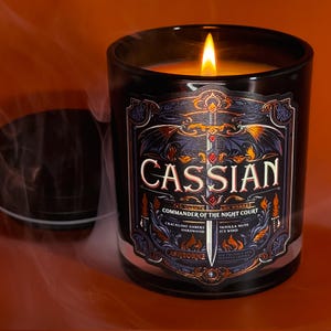 May include: A lit black glass candle with a label that reads "CASSIAN, Commander of the Night Court." The label features a sword, wings, and decorative elements. The candle's scent is "Crackling Embers, Hardwood, Vanilla Musk, Icy Wind."