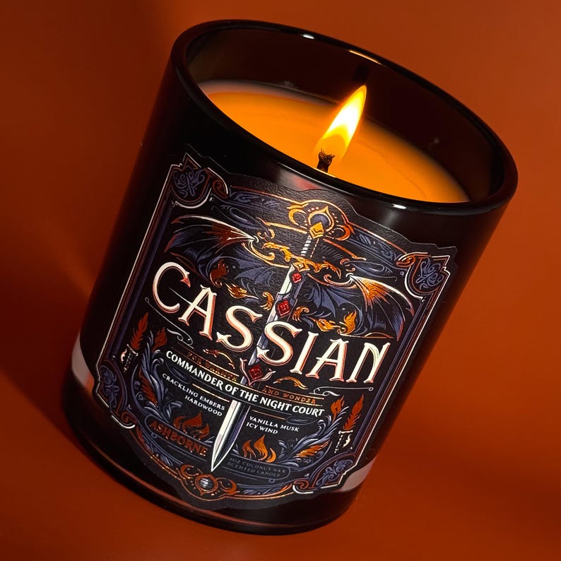 May include: A lit black candle with a label that reads "CASSIAN Commander of the Night Court." The label features a sword and dragon design with the words "Crackling Embers Hardwood" and "Vanilla Musk Icy Wind." The candle is set against an orange background.