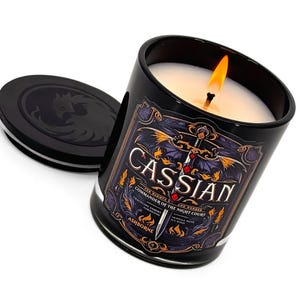 May include: A lit black candle with a label that reads "Cassian Commander of the Night Court". The candle is in a black glass jar with a matching lid. The label features a sword and dragon design. The candle is labeled "Ashborne".