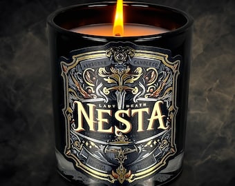 Puede incluir: Una vela negra encendida en un tarro de vidrio con una etiqueta decorativa. La etiqueta presenta el texto "LADY DEATH NESTA" en letras doradas, con adornos dorados y plateados. La vela es de Ashborne Candle Co.