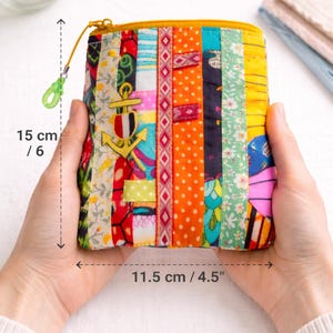 Può includere: Un astuccio con cerniera patchwork colorato, largo 11,5 cm e alto 15 cm, tenuto in mano. L'astuccio presenta strisce verticali di tessuto fantasia in vari colori, tra cui arancione, giallo e blu. È visibile un ciondolo a forma di ancora.