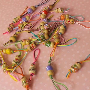 Puede incluir: Una colección de pequeños colgantes de cuentas de madera con diseños pintados y cuerdas. Las cuentas presentan varios patrones, como puntos y cruces, en tonos rojos, verdes, morados y amarillos. Las cuerdas son de varios colores.