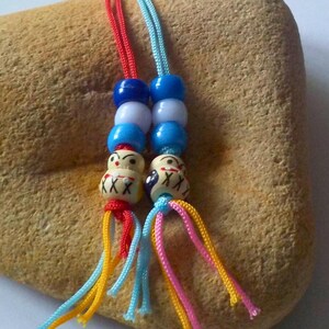 Può includere: Due ciondoli decorativi con perline di plastica impilate in blu, bianco e blu scuro, che terminano in piccole perline rotonde color crema dipinte con tratti del viso neri e rossi. Ogni ciondolo pende da un cordino con fili multicolori.