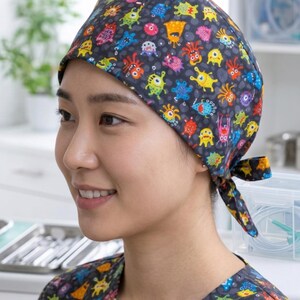 Puede incluir: Un gorro quirúrgico gris oscuro con un patrón colorido de monstruos de dibujos animados. El gorro está atado en la parte posterior y lo lleva una persona. La persona lleva una parte superior de uniforme a juego.