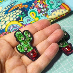 Peut inclure: Deux écussons brodés en forme de cactus avec du fil vert, des accents en cristal transparent et une base de pot rouge foncé ornée d'un grand cristal rouge. Les écussons ont une bordure noire cousue pour une application sur tissu.
