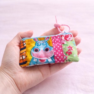 Può includere: Un piccolo portamonete patchwork colorato con un ippopotamo dei cartoni animati. Il portamonete ha una cerniera blu e un cordino rosa. Il patchwork include motivi arancioni, gialli, pois rosa e floreali.