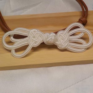 Puede incluir: Un collar de nudo de seda blanca con un cordón de cuero marrón. El nudo está intrincadamente diseñado con múltiples bucles y un nudo central. El collar se exhibe sobre una superficie de madera clara.