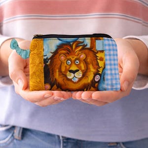 May include: A small, rectangular zipped pouch with a cartoon lion design. The pouch has a blue and white gingham pattern on one end, a yellow floral pattern on the other, and a blue fabric pull.