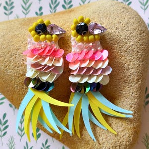 Peut inclure: Une paire de boucles d'oreilles en forme d'oiseau ornées de perles de rocaille jaunes, de cristaux noirs et transparents, et de sequins superposés roses, pêche et blancs. Les queues sont faites de fines bandes bleu clair et jaunes.