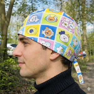 Peut inclure: Un bonnet de chirurgien coloré avec un motif répété de personnages de dessins animés. Le bonnet est noué dans le dos et est porté par une personne. L'arrière-plan est flou, avec des arbres et du feuillage visibles.