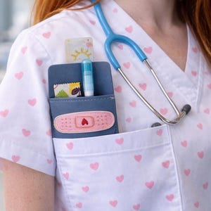 Peut inclure: Haut de blouse médicale blanc avec des cœurs roses, un stéthoscope bleu et un organiseur de poche bleu. L'organisateur contient un pansement, une petite carte et un stylo bleu. Le haut a un col en V et une poche poitrine.