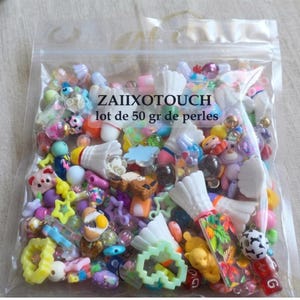 Peut inclure: Un sachet en plastique transparent contenant un assortiment de 50 g de perles et breloques multicolores de formes variées, incluant des volants blancs, des étoiles, des animaux et des cubes lettres, avec le texte ZAIIXOTOUCH lot de 50 gr de perles.