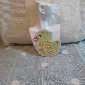 Peut inclure: Un patch brodé vert représentant un canard avec des fleurs orange et roses. Le canard est assis sur une surface blanche.