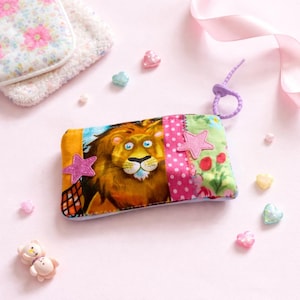 Può includere: Un astuccio rettangolare in tessuto colorato con un'illustrazione di un leone dei cartoni animati. L'astuccio ha un anello viola ed è decorato con stelle rosa e motivi floreali. Piccoli abbellimenti a forma di cuore e stella circondano l'astuccio.