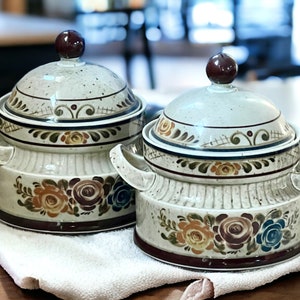 May include: Two ceramic serving dishes with lids. The dishes are white with brown, orange, and blue floral designs. The lids have a brown knob.