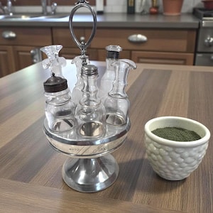 May include: A silver condiment set with six clear glass bottles and a white textured bowl filled with green herbs. The set has a handle and sits on a wooden surface, perfect for kitchen or dining decor.