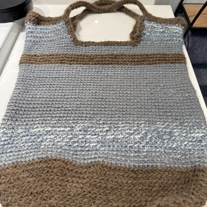 May include: A hand-knitted tote bag in shades of gray, brown, and blue. The bag features a textured, woven design with brown handles and a brown bottom. The bag is rectangular with a rounded bottom.