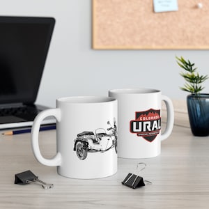 May include: Two white ceramic mugs with black and red designs. One mug features a black and white illustration of a motorcycle with a sidecar. The other mug has a red and black logo that says "Colorado Ural Sidecar Motorcycles".