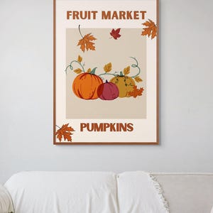 Peut inclure: Une affiche encadrée avec les mots "FRUIT MARKET" et "PUMPKINS". L'illustration représente trois citrouilles orange, rouge et jaune, avec des feuilles d'automne et des vignes. Le fond est beige neutre.