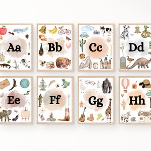 May include: Eight printable alphabet posters with watercolor illustrations of objects starting with each letter. The posters feature a light brown background with a peach-colored watercolor cloud in the center. Each poster has the letter in black text and the letter name in black text below.