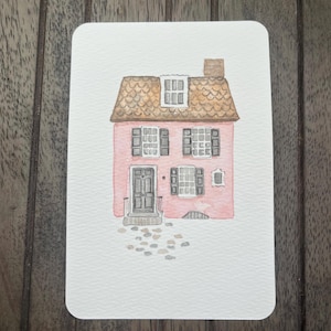 May include: Watercolor painting of a pink house with a brown roof and a chimney. The house has multiple windows with black shutters and a front door. A small stone path leads to the door. The artwork is on a white rectangular card.