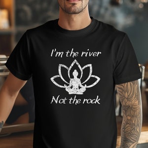 Puede incluir: Una camiseta negra con un gráfico blanco de una flor de loto y una persona sentada en meditación. El texto "I'm the river Not the rock" está impreso debajo del gráfico.