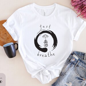 May include: White t-shirt with a black and white graphic design. The graphic features a circle with a lotus flower and the text "just breathe".