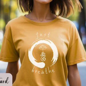 May include: A mustard yellow t-shirt with a white graphic design. The graphic features a circle with a lotus flower and the text "just breathe".
