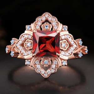 May include: Rose gold ring featuring a square, deep red gemstone at the center, surrounded by smaller white and blue gemstones. The ring has an ornate, floral-inspired design with intricate details, perfect for a special occasion.