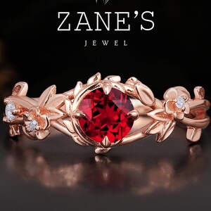 May include: Rose gold ring with a central, round, red gemstone, encircled by a floral design and small diamonds. The ring is from Zane's Jewel.