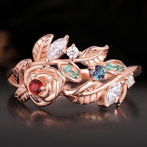 May include: Rose gold ring featuring a rose and leaf design. The ring is adorned with a red gemstone at the center of the rose, along with white, green, and blue gemstones. The ring is set against a dark background.