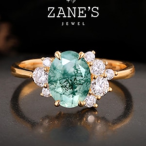 Op de afbeelding: Een gouden ring met een grote, ovaal geslepen, lichtgroene edelsteen met interne patronen, omringd door kleinere, heldere edelstenen. De ring heeft een delicate band en de tekst "Zane's Jewel" in een diamantlogo.