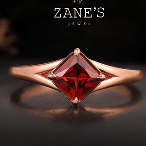 May include: A rose gold ring with a square, deep red gemstone. The ring has a delicate band and a setting that highlights the gemstone. The image includes the text "Zane's Jewel".