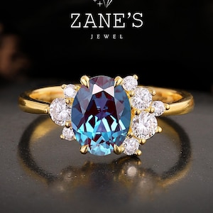 Op de afbeelding: Een gouden ring met een ovale alexandriet edelsteen, die van kleur verandert, omringd door kleine, ronde diamanten. De ring heeft een gepolijste gouden band. De tekst "Zane's Jewel" staat boven de ring.