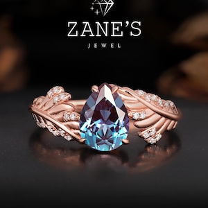 Op de afbeelding: Een roségouden ring met een peervormige, blauw-paarse edelsteen. De ring heeft een band met bladvormige details en kleine, sprankelende accentstenen. De tekst "ZANE'S JEWEL" is zichtbaar.
